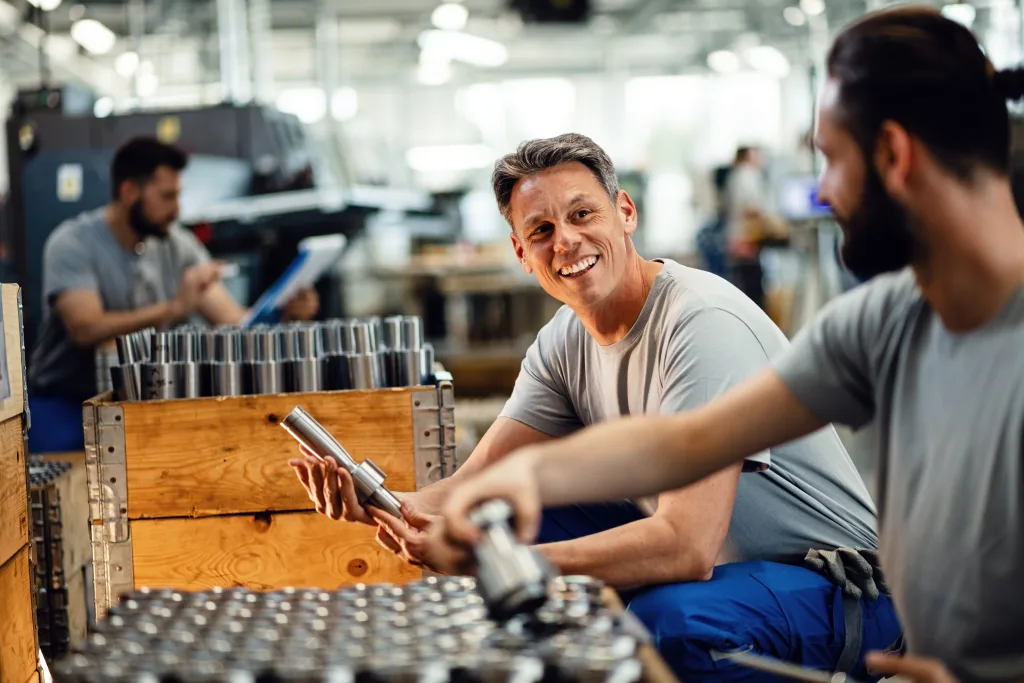 Zwei glückliche Stahlarbeiter, die Produkte für den Vertrieb vorbereiten und in einer Industrieanlage kommunizieren. Der Fokus liegt auf dem mittleren erwachsenen Arbeiter.